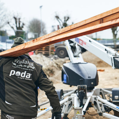 Monteur draagt lange houten planken op een bouwplaats met hoogwerker en materialen.