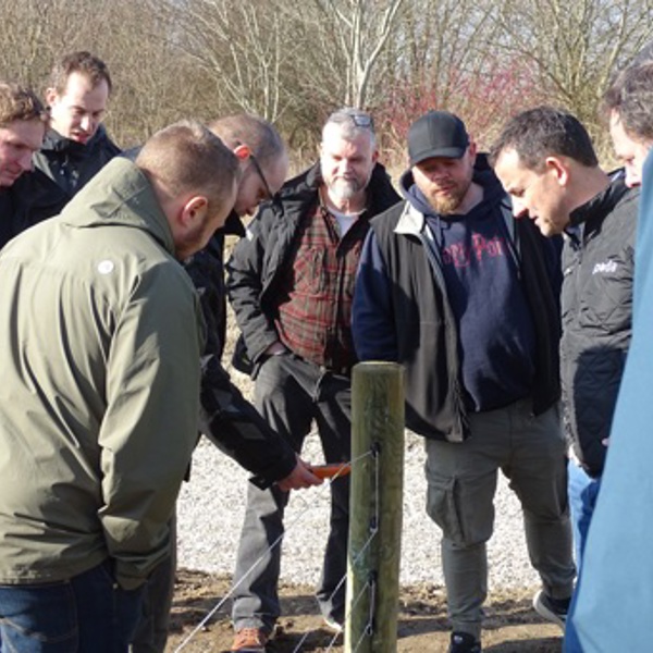 Poda Partners verzamelen zich rond een afrasteringspaal tijdens een training om te leren hoe je een spanningsmeter voor elektrische afrasteringen gebruikt.