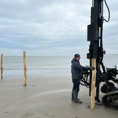 Monteur plaatst houten palen met een machine op een strand aan de kust.