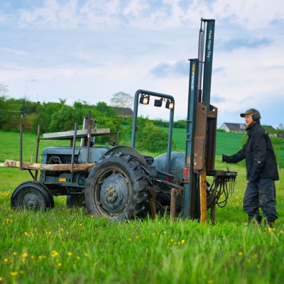 Monteur werkt met een palenrammer op een tractor bij het plaatsen van omheiningspalen in een groen veldgebied.