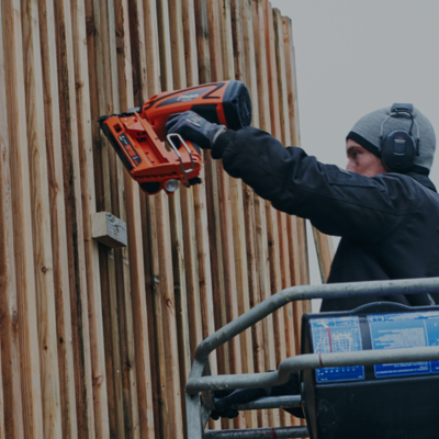 Monteur bevestigt houten bekleding aan een hoge houten omheining vanuit een hoogwerker.